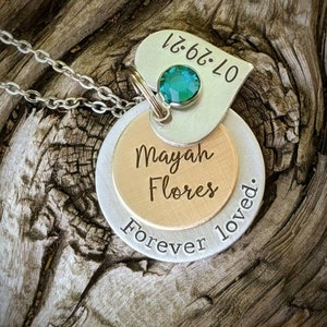 May include: A silver necklace with three layered pendants. The top pendant is heart-shaped with the date 07.29.21 and a green gemstone. The middle pendant is gold-colored with the name "Mayah Flores". The bottom pendant says "Forever loved."