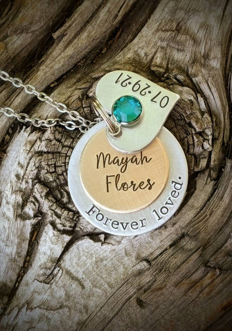 May include: A silver necklace with three layered pendants. The top pendant is heart-shaped with the date 07.29.21 and a green gemstone. The middle pendant is gold-colored with the name "Mayah Flores". The bottom pendant says "Forever loved."