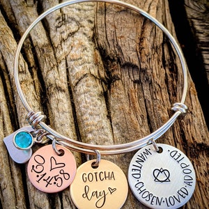 May include: A silver bangle bracelet with three charms. The charms are round and have the following text: "Gotcha Day", "1:45:30", and "Adopted, Loved, Chosen, Wanted".