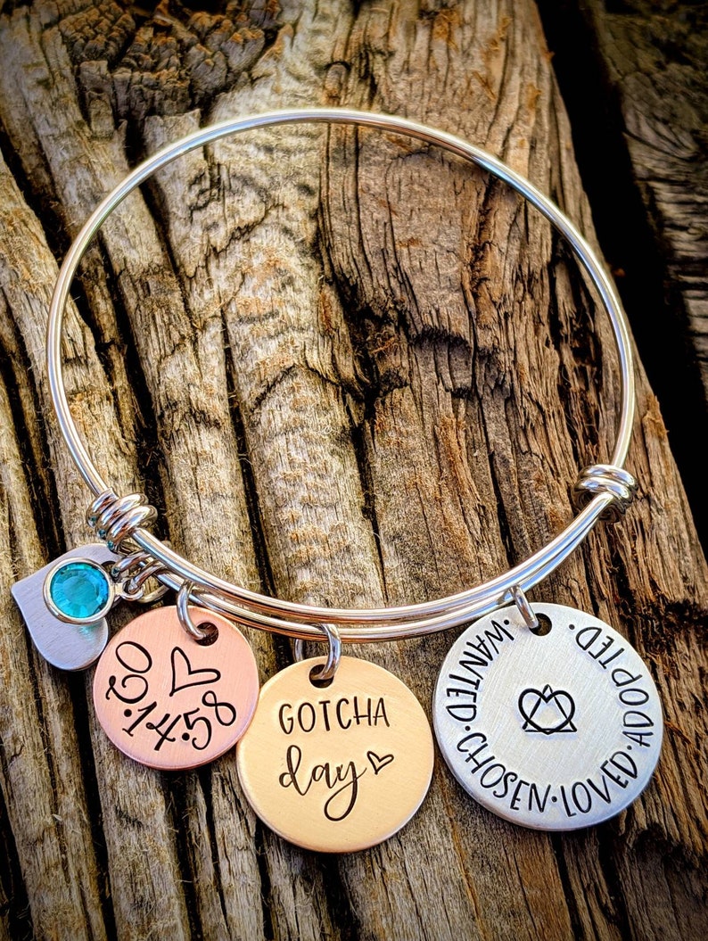 May include: A silver bangle bracelet with three charms. The charms are round and have the following text: "Gotcha Day", "1:45:30", and "Adopted, Loved, Chosen, Wanted".