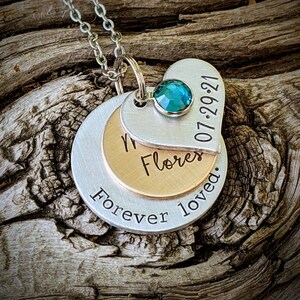 May include: A silver necklace with three layered pendants. The top pendant is heart-shaped with a teal gemstone and the date "07-29-21". The middle pendant is gold-colored, and the bottom pendant is silver with the words "Forever loved."