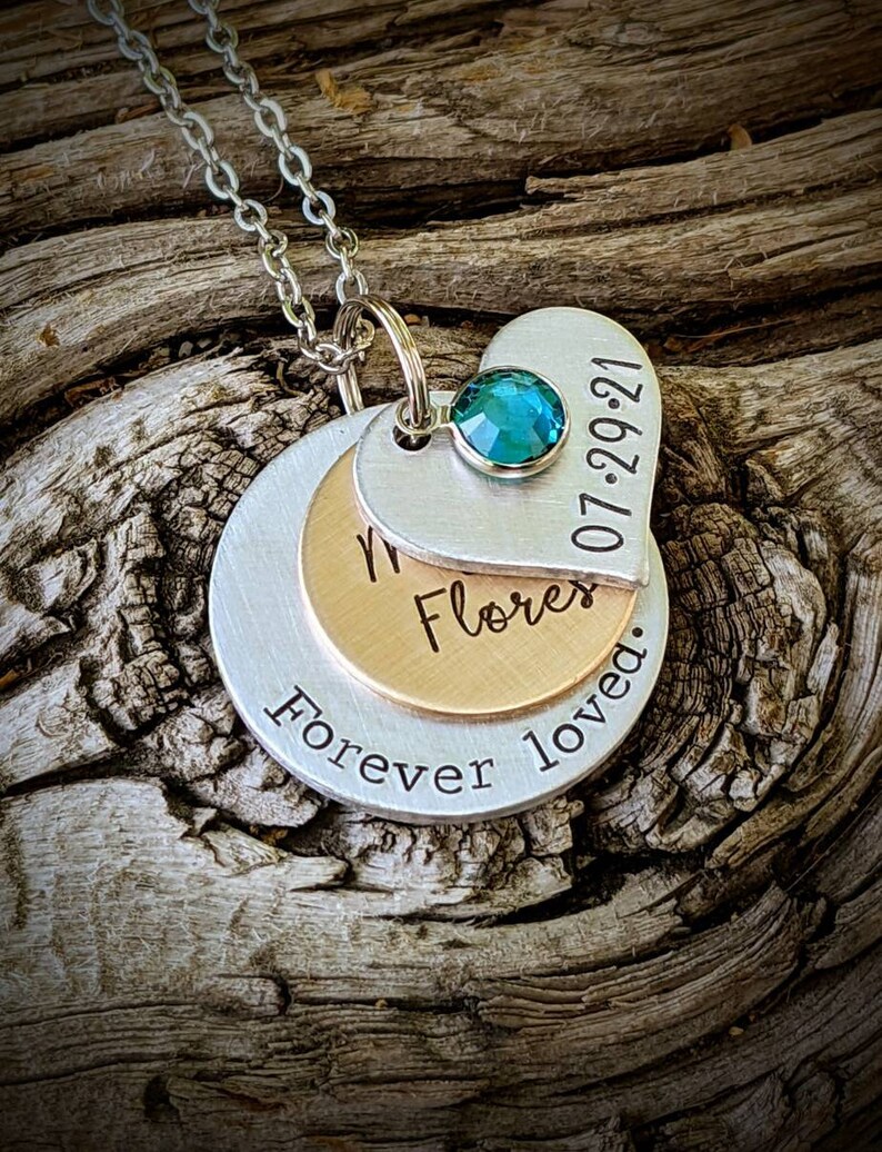 May include: A silver necklace with three layered pendants. The top pendant is heart-shaped with a teal gemstone and the date "07-29-21". The middle pendant is gold-colored, and the bottom pendant is silver with the words "Forever loved."