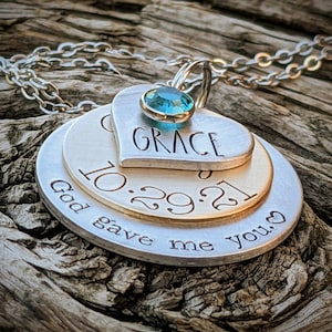May include: A silver necklace with three layered charms. The top charm is a heart with the word "GRACE" inscribed on it. The middle charm is a round disc with the date "10.29.21" inscribed on it. The bottom charm is a round disc with the text "God gave me you." inscribed on it. A light blue gemstone is attached to the top charm.