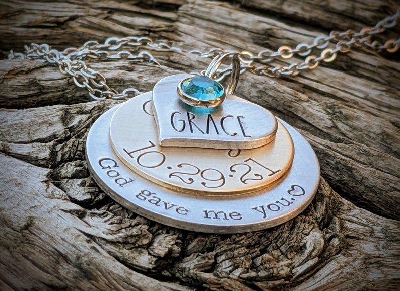 May include: A silver necklace with three layered charms. The top charm is a heart with the word "GRACE" inscribed on it. The middle charm is a round disc with the date "10.29.21" inscribed on it. The bottom charm is a round disc with the text "God gave me you." inscribed on it. A light blue gemstone is attached to the top charm.