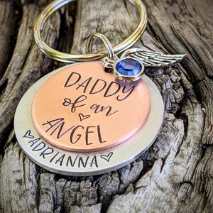 May include: A silver and copper keychain with a blue gem and angel wings charm. The keychain says "Daddy of an Angel" and "Adrianna" with a heart symbol.