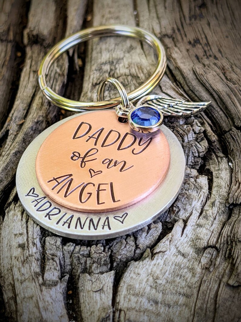May include: A silver and copper keychain with a blue gem and angel wings charm. The keychain says "Daddy of an Angel" and "Adrianna" with a heart symbol.