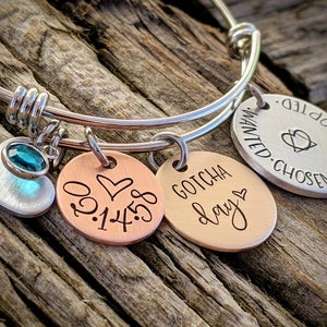 May include: A silver bangle bracelet with three charms. The charms are a silver heart with a teal gemstone, a copper charm with the date 01.14.18 and a heart, and a copper charm with the text "GOTCHA day" and a heart.