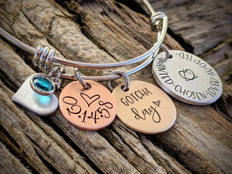 May include: A silver bangle bracelet with three charms. The charms are a silver heart with a teal gemstone, a copper charm with the date 01.14.18 and a heart, and a copper charm with the text "GOTCHA day" and a heart.