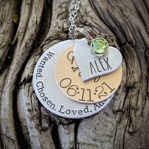 May include: Silver necklace with three layered discs. The top disc is a heart with the name "ALEX" engraved on it. The middle disc is gold and has the date "06.11.21" engraved on it. The bottom disc is silver and has the text "Wanted. Chosen. Loved. Adored" engraved on it. A green gemstone is attached to the top of the heart.
