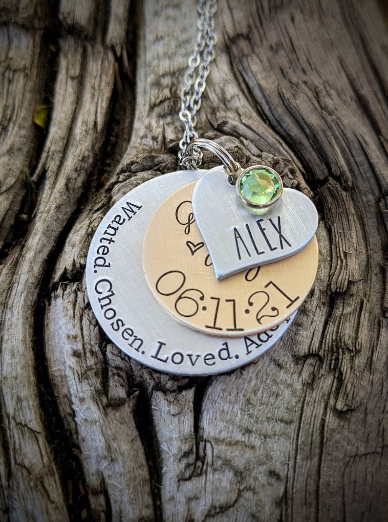 May include: Silver necklace with three layered discs. The top disc is a heart with the name "ALEX" engraved on it. The middle disc is gold and has the date "06.11.21" engraved on it. The bottom disc is silver and has the text "Wanted. Chosen. Loved. Adored" engraved on it. A green gemstone is attached to the top of the heart.