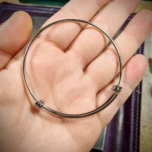 May include: A silver bangle bracelet with two round clasps. The bracelet is made of thin metal and is adjustable.