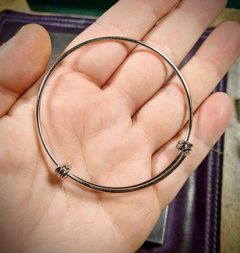 May include: A silver bangle bracelet with two round clasps. The bracelet is made of thin metal and is adjustable.