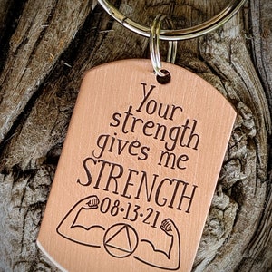 May include: A copper-colored dog tag keychain with the words "Your strength gives me STRENGTH" engraved in black. The tag also features a date and arm muscle illustrations. The keychain is attached to a silver ring.