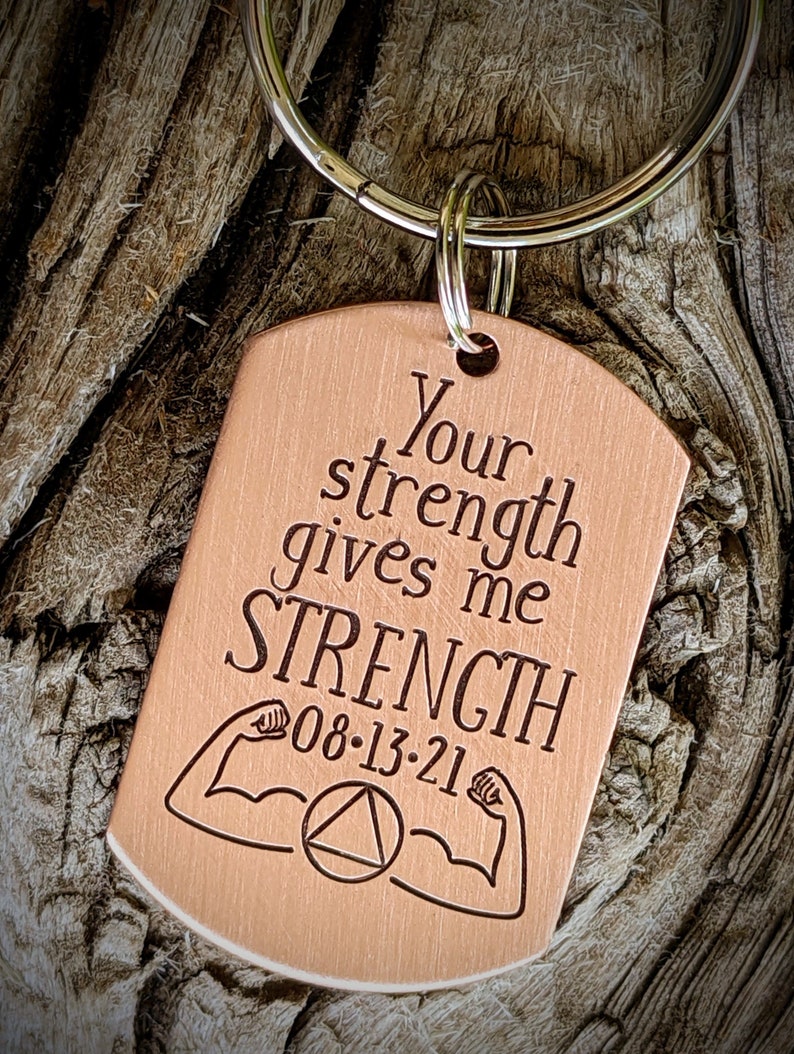 May include: A copper-colored dog tag keychain with the words "Your strength gives me STRENGTH" engraved in black. The tag also features a date and arm muscle illustrations. The keychain is attached to a silver ring.