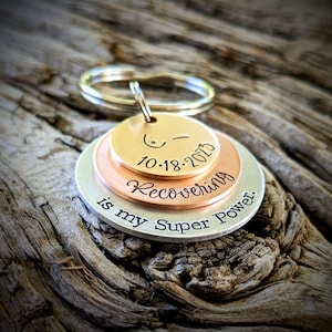 May include: A silver, copper, and gold layered keychain with the text "Recovering is my Super Power." and the date "10-18-2013".