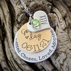 May include: A silver necklace with a heart charm engraved with the name "Alex" and a green gemstone. The heart charm is attached to a round charm with the text "Go day 06.11.21 Wanted. Chosen. Loved. Adopted."