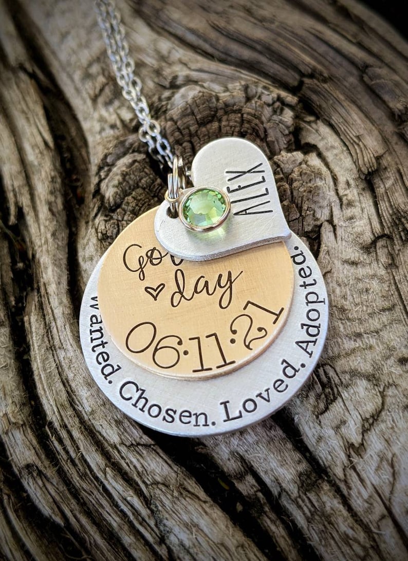 May include: A silver necklace with a heart charm engraved with the name "Alex" and a green gemstone. The heart charm is attached to a round charm with the text "Go day 06.11.21 Wanted. Chosen. Loved. Adopted."