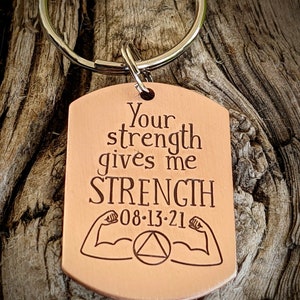 May include: Copper-colored dog tag keychain with the words "Your strength gives me STRENGTH" engraved in a serif font. The tag includes the date "08-13-21", flexing arm graphics, and a triangle symbol. A silver-colored keyring is attached.