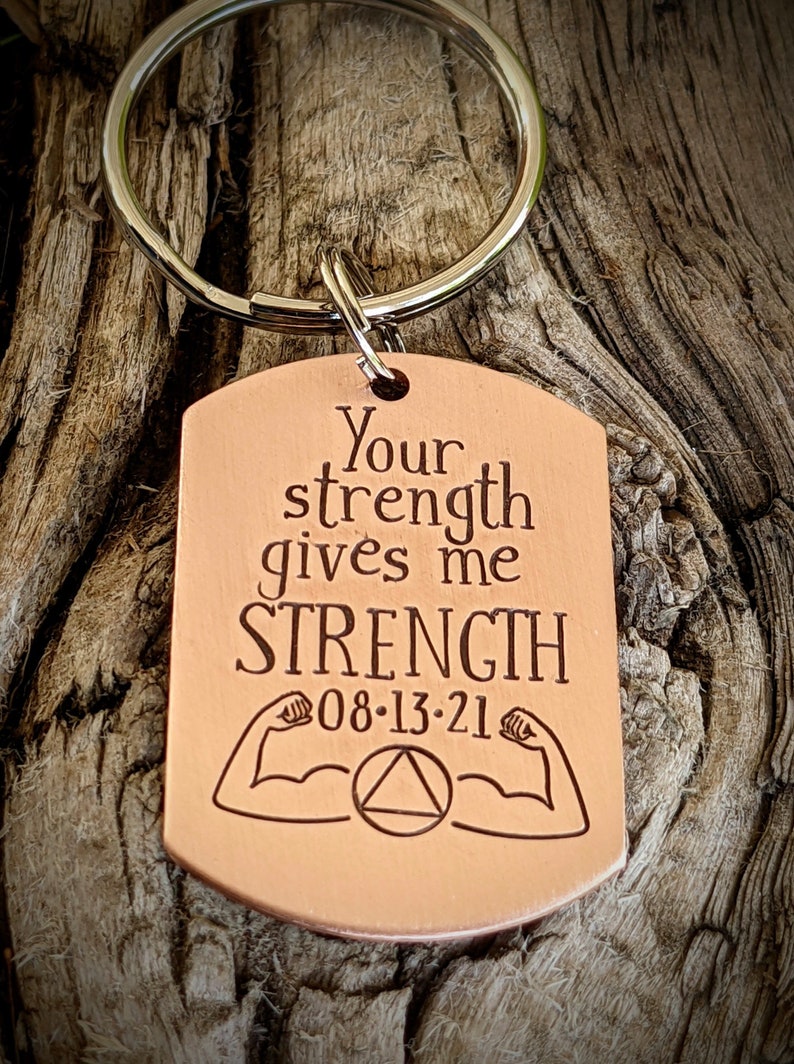 May include: Copper-colored dog tag keychain with the words "Your strength gives me STRENGTH" engraved in a serif font. The tag includes the date "08-13-21", flexing arm graphics, and a triangle symbol. A silver-colored keyring is attached.