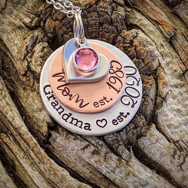 May include: Silver necklace with a layered pendant featuring the words "Mom est. 1989" and "Grandma est. 2009" with a heart symbol between the words. The pendant has a pink rhinestone in the center.
