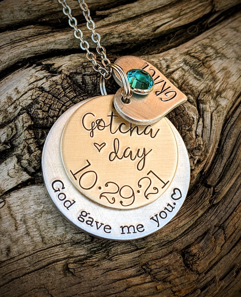 May include: A silver necklace with a heart charm that says "GRACE" and a round charm that says "Gotcha day 10.29.21 God gave me you." The heart charm has a teal blue gemstone.