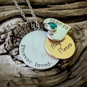 May include: A silver necklace with multiple pendants. The largest pendant is a silver disc with the words "Forever loved." A smaller gold disc is engraved with "Megan Flores." A heart-shaped pendant with a teal gemstone and the date "07-29-21" is also included.