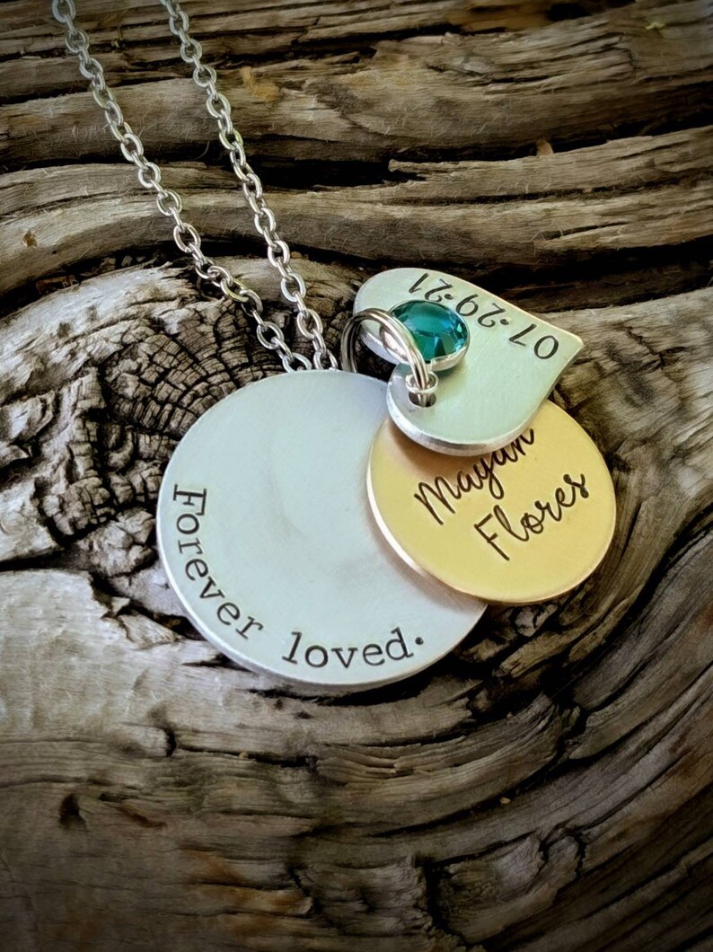 May include: A silver necklace with multiple pendants. The largest pendant is a silver disc with the words "Forever loved." A smaller gold disc is engraved with "Megan Flores." A heart-shaped pendant with a teal gemstone and the date "07-29-21" is also included.