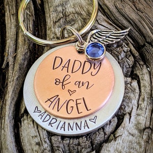 May include: A silver and copper keychain with a blue crystal and a silver angel wing charm. The keychain has the text "Daddy of an Angel" and "Adrianna" engraved on it.