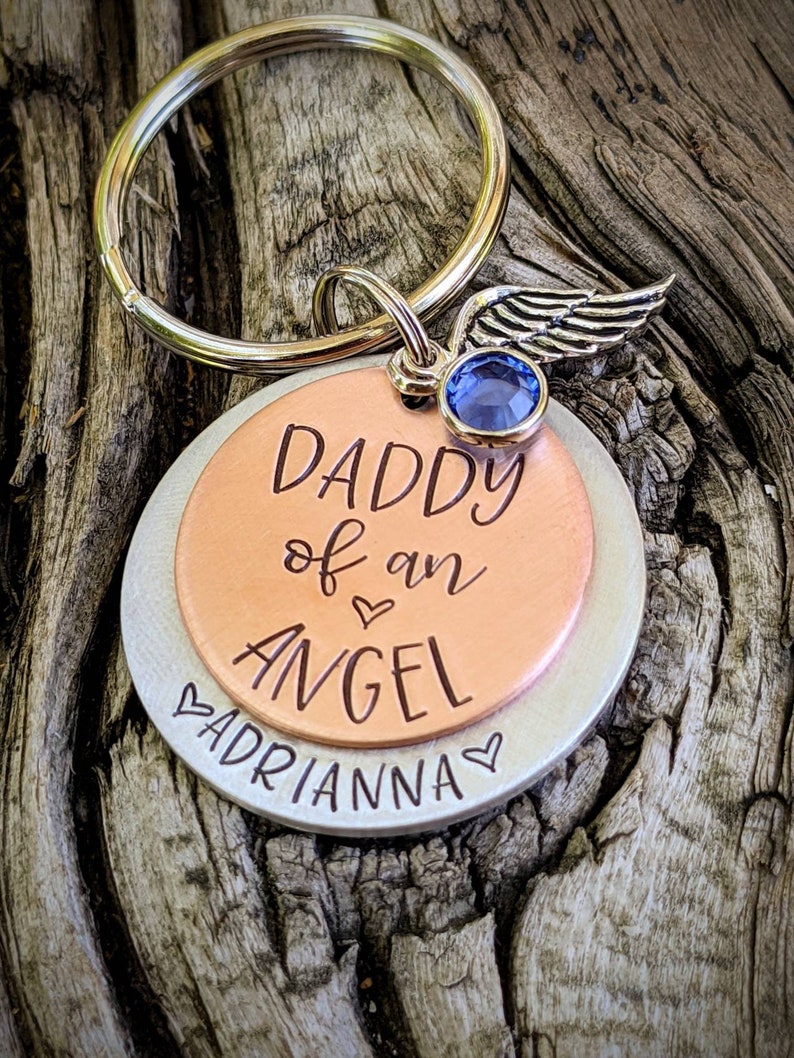 May include: A silver and copper keychain with a blue crystal and a silver angel wing charm. The keychain has the text "Daddy of an Angel" and "Adrianna" engraved on it.