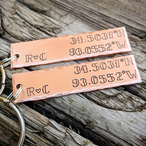 May include: Two copper keychains with a hammered finish. Each keychain has a small heart and the initials "R & C" engraved on one side. The other side has the coordinates "34.5037°N 93.0552°W" engraved on it.