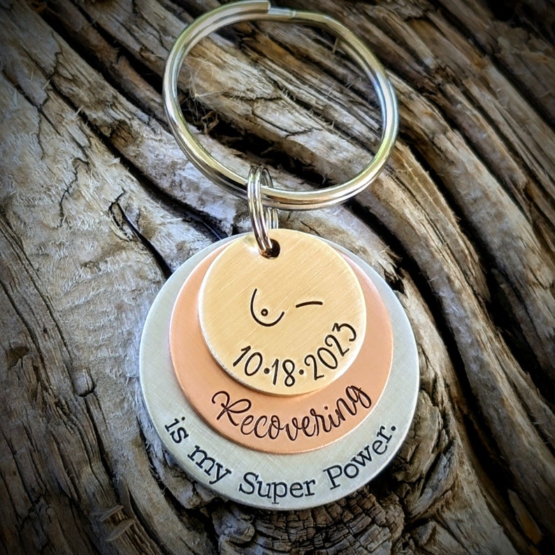 May include: A silver, copper, and rose gold layered keychain with the text "10-18-2023 Recovering is my Super Power."