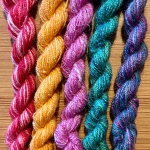 May include: Five twisted skeins of yarn arranged vertically. The colours from left to right are a variegated red and white, a solid golden orange, a variegated magenta and light pink, a variegated teal and green, and a variegated blue and purple.