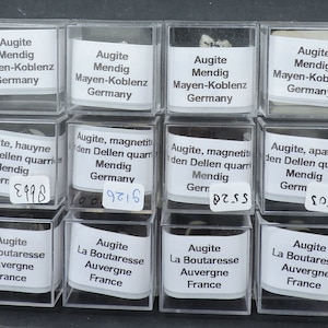 May include: A collection of 18 small, clear plastic boxes, each containing a different type of mineral specimen. The boxes are arranged in three rows of six. Each box has a white label with the name of the mineral, the location where it was found, and a number. The minerals include augite, hauyne, magnetite, apatite, and titanite.