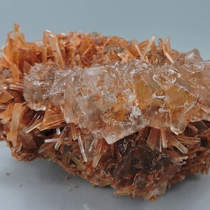 Puede incluir: Un grupo de cristales transparentes y vítreos con un tono marrón rojizo, dispuestos en una matriz de cristales más pequeños en forma de aguja. Los cristales más grandes tienen una forma cúbica, mientras que los más pequeños son alargados y puntiagudos.