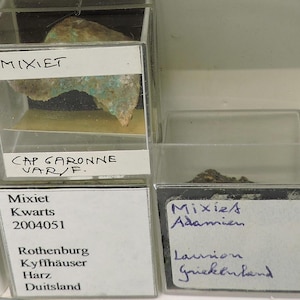 May include: A collection of mineral specimens displayed in clear plastic boxes. The top box contains a piece of mixiet with green and brown hues. The labels include the text "MIXIET" and "CAP GARONNE VAR/F."