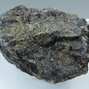 May include: A dark gray rock with green and black mineral inclusions. The rock has a rough, uneven surface.