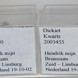May include: Two small, clear plastic display cases, each containing a small, gray mineral specimen. The cases have white labels with black text, including the words "Dickiet", "Kwarts", and a series of numbers and text.