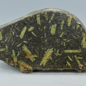Op de afbeelding: Een ruwe, onregelmatig gevormde steen met een donkergrijze basiskleur. De steen is sterk bespikkeld met lichtgroene, langwerpige kristalformaties. Het geheel doet denken aan een natuurlijk, geologisch specimen.