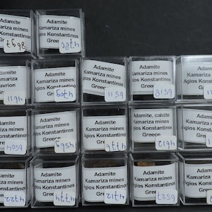 May include: Twenty small clear plastic boxes with white labels containing Adamite mineral specimens from the Kamariza mines in Agios Konstantinos, Greece. The labels include the mineral name, location, and a number.