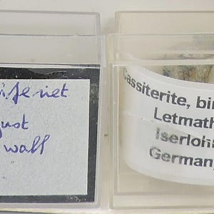 Puede incluir: Dos pequeñas vitrinas de plástico transparente. La caja izquierda tiene un fondo negro con texto manuscrito: "Kassiteriet, St. Just, Cornwall, G.B." La caja derecha tiene una muestra mineral y una etiqueta con texto: "Cassiterita, bindheim, Letmathe, Iserlohn, Alemania."