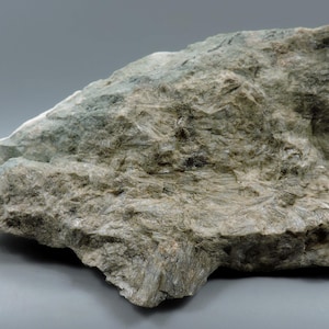 May include: A large, irregularly shaped rock formation with a rough, textured surface. The rock displays a mix of gray, green, and tan hues, with visible crystalline structures. The overall appearance suggests a natural mineral specimen.