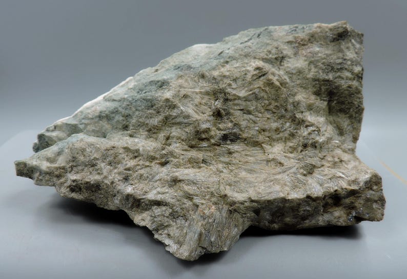 May include: A large, irregularly shaped rock formation with a rough, textured surface. The rock displays a mix of gray, green, and tan hues, with visible crystalline structures. The overall appearance suggests a natural mineral specimen.