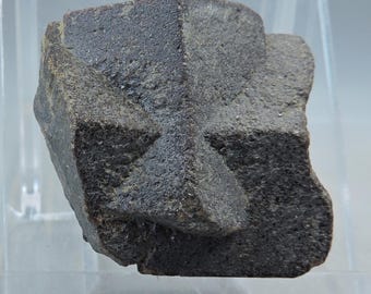 May include: A dark gray mineral specimen with a cross-shaped formation. The stone has a rough, textured surface and appears to be a natural, unpolished rock. The lighting highlights the angular shapes and the subtle variations in color.