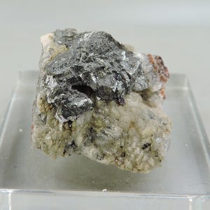 Può includere: Un campione minerale con un mix di colori e trame. La parte superiore è di un grigio scuro metallico, mentre la base è beige chiaro e granulare. Sono visibili anche piccole macchie di marrone rossastro. Il minerale poggia su una base rettangolare trasparente.