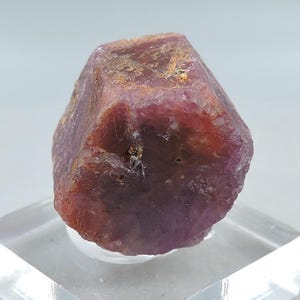 Könnte beinhalten: Ein roher, sechseckiger Rubinkristall mit einem tiefen rot-violetten Farbton und einer texturierten, undurchsichtigen Oberfläche. Der Edelstein weist natürliche mineralische Einschlüsse und eine facettierte, geometrische Struktur auf.