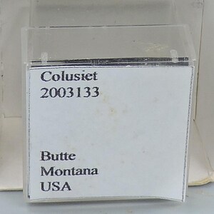 Może przedstawiać: Biała etykieta w przezroczystej plastikowej obudowie. Etykieta zawiera tekst "Colusiet" nad numerem "2003133". Poniżej znajduje się tekst "Butte Montana USA".