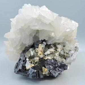 May include: A natural mineral specimen featuring a cluster of white, crystalline formations atop a base of dark, metallic-looking minerals. The specimen has a rough, textured appearance with hints of yellow and green.