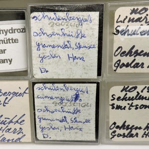 May include: A collection of labeled mineral samples in clear plastic cases. The labels, handwritten in blue ink, identify the minerals as "Schulenbergit" and "Linarite" from Ochsenhütte, Goslar, Germany. One label reads "Smitsonit".