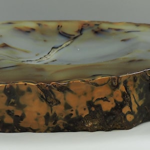 Op de afbeelding: Een grote, natuurlijke agaat geode kom met een lichtgrijze binnenkant en bruine, witte en zwarte banden aan de buitenkant. De kom is onregelmatig gevormt en heeft een ruwe, natuurlijke textuur.