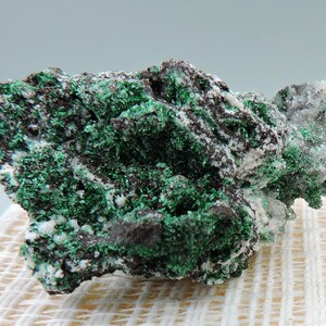 Può includere: Un esemplare minerale grezzo caratterizzato da un ammasso di strutture cristalline verde smeraldo vibrante intervallate da una matrice rocciosa marrone scuro e bianca. La superficie della pietra presenta un aspetto testurizzato e frastagliato.